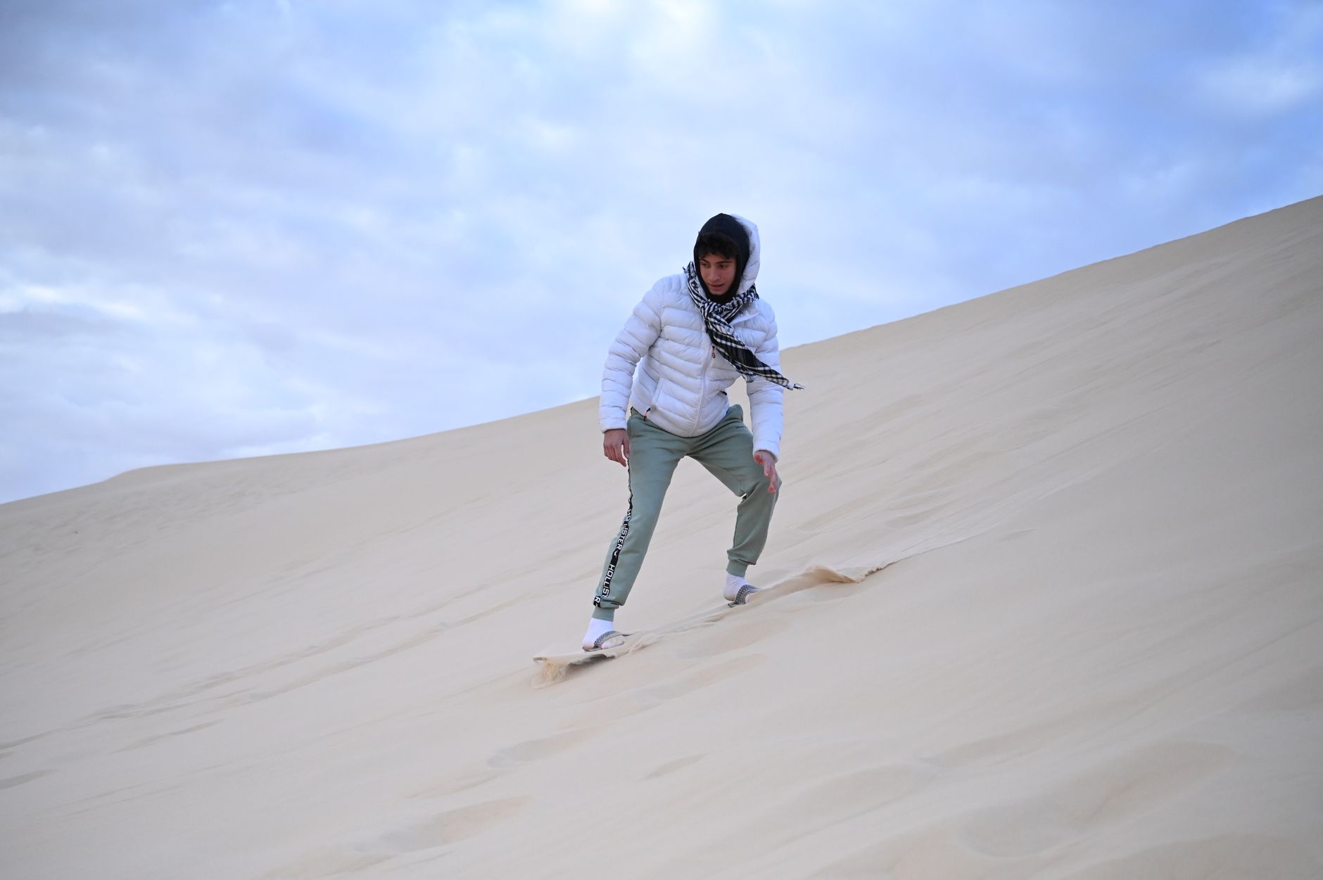 A man in a white jacket sandboarding on the vast desert sand dunes under a cloudy sky.