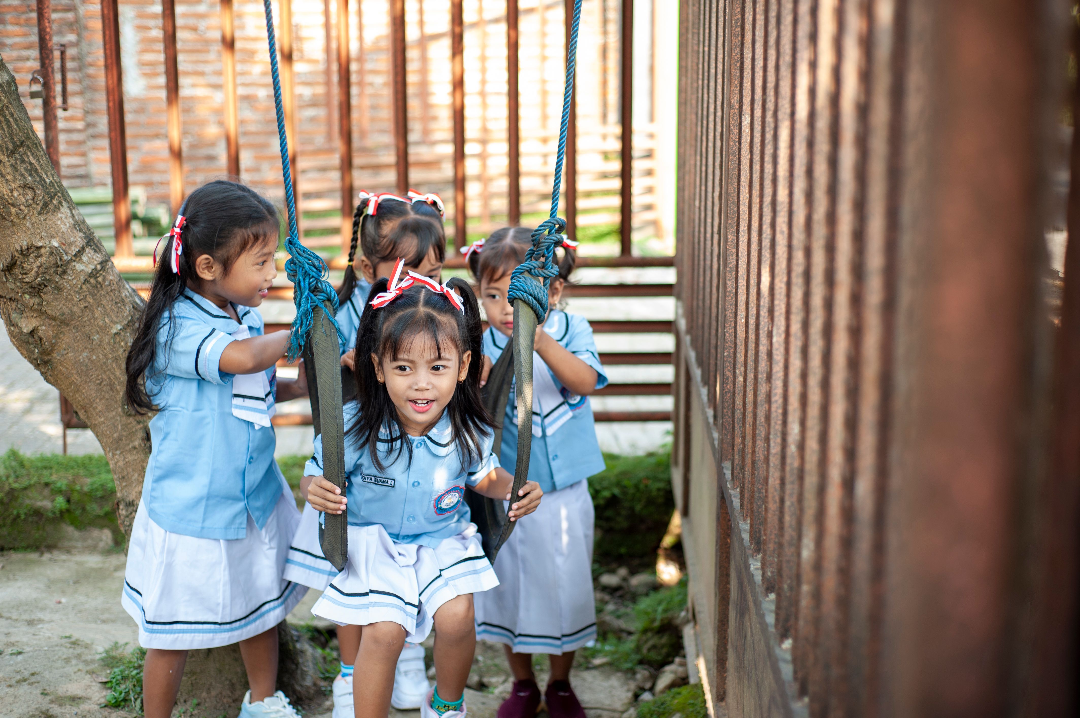 Four young girls in blue uniforms enjoying playtime on a swing set outdoors.