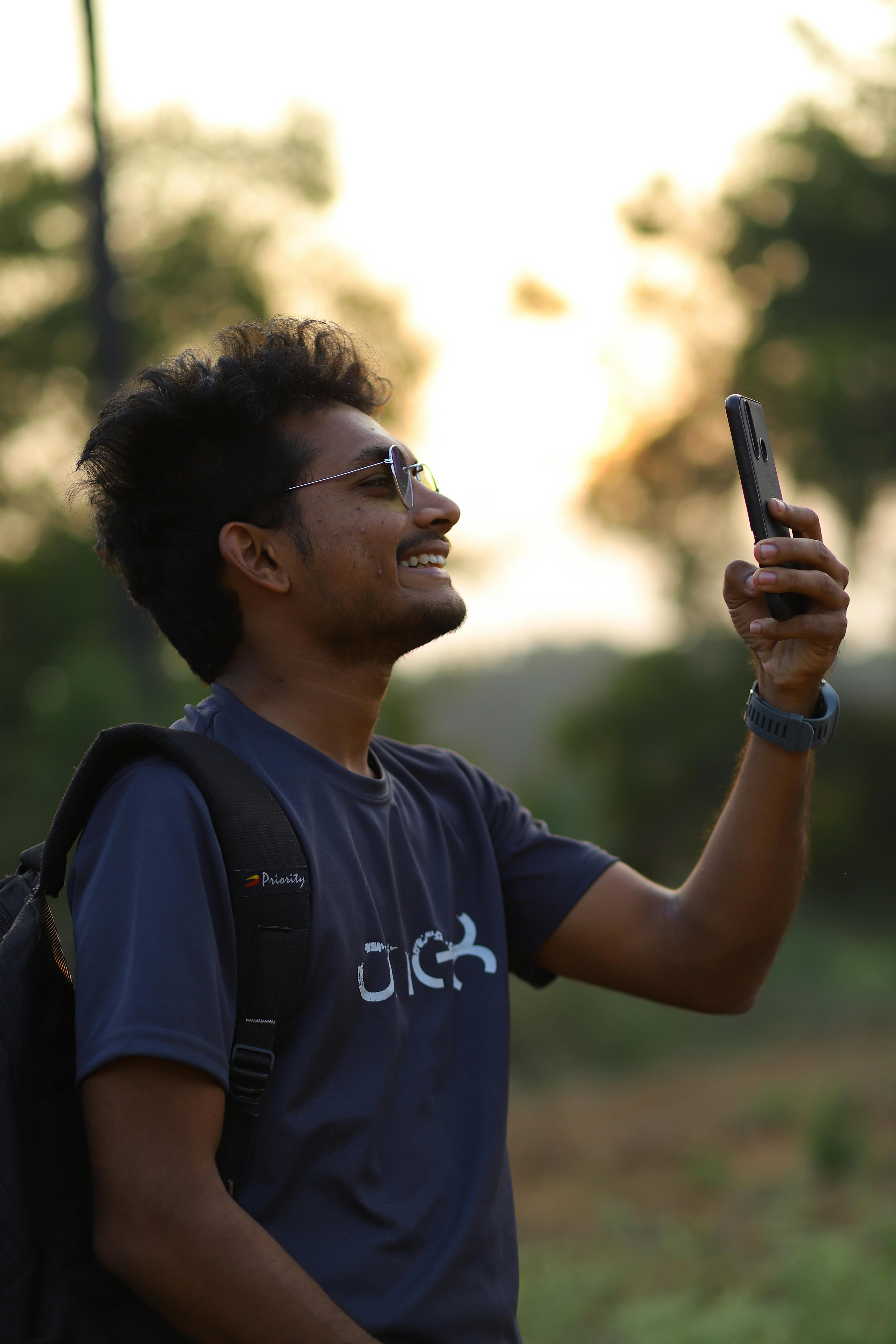 Casual man with backpack smiling and photographing with smartphone at sunset outdoors.