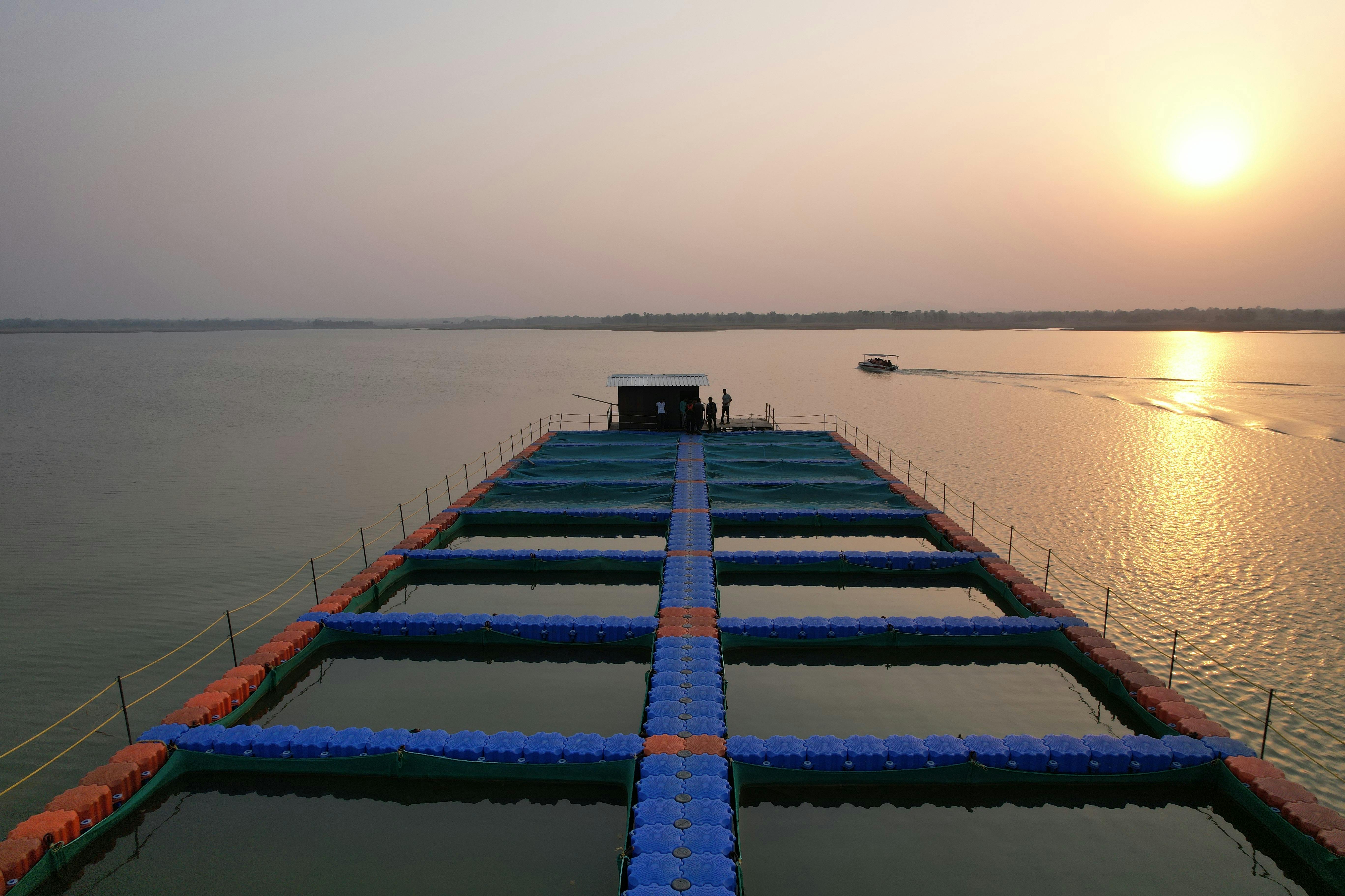 A tranquil aerial view of a floating fish farm at sunset with a boat crossing calm lake waters.