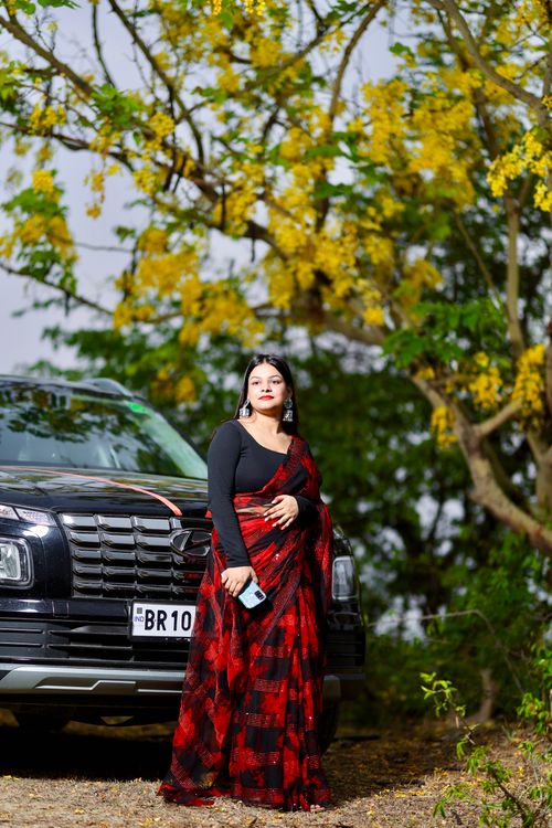 Free A woman in a red sari stands beside a black car outdoors among yellow flowering trees. Stock Photo