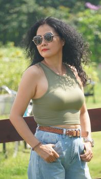 Fashionable woman with curly hair and sunglasses standing outdoors in nature