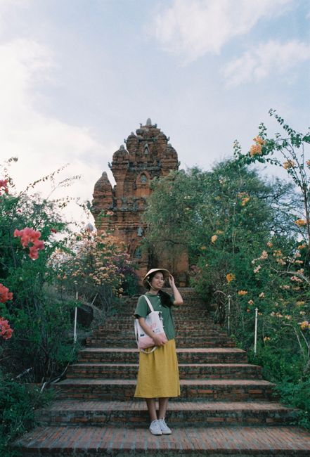 Woman poses in front of a historic temple amid vibrant greenery and flowers.