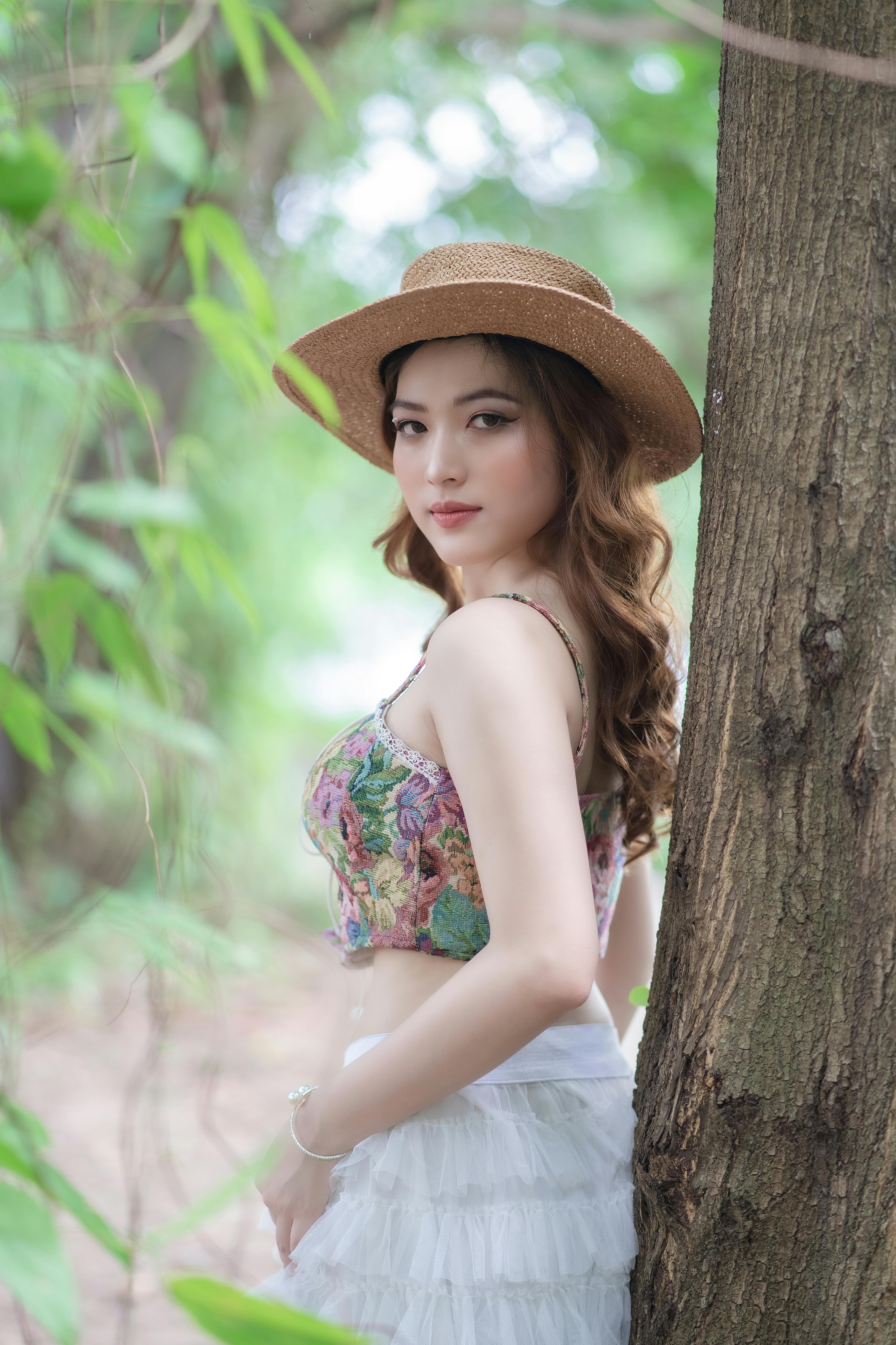 Stylish brunette woman in a summer outfit and straw hat standing by a tree outdoors.
