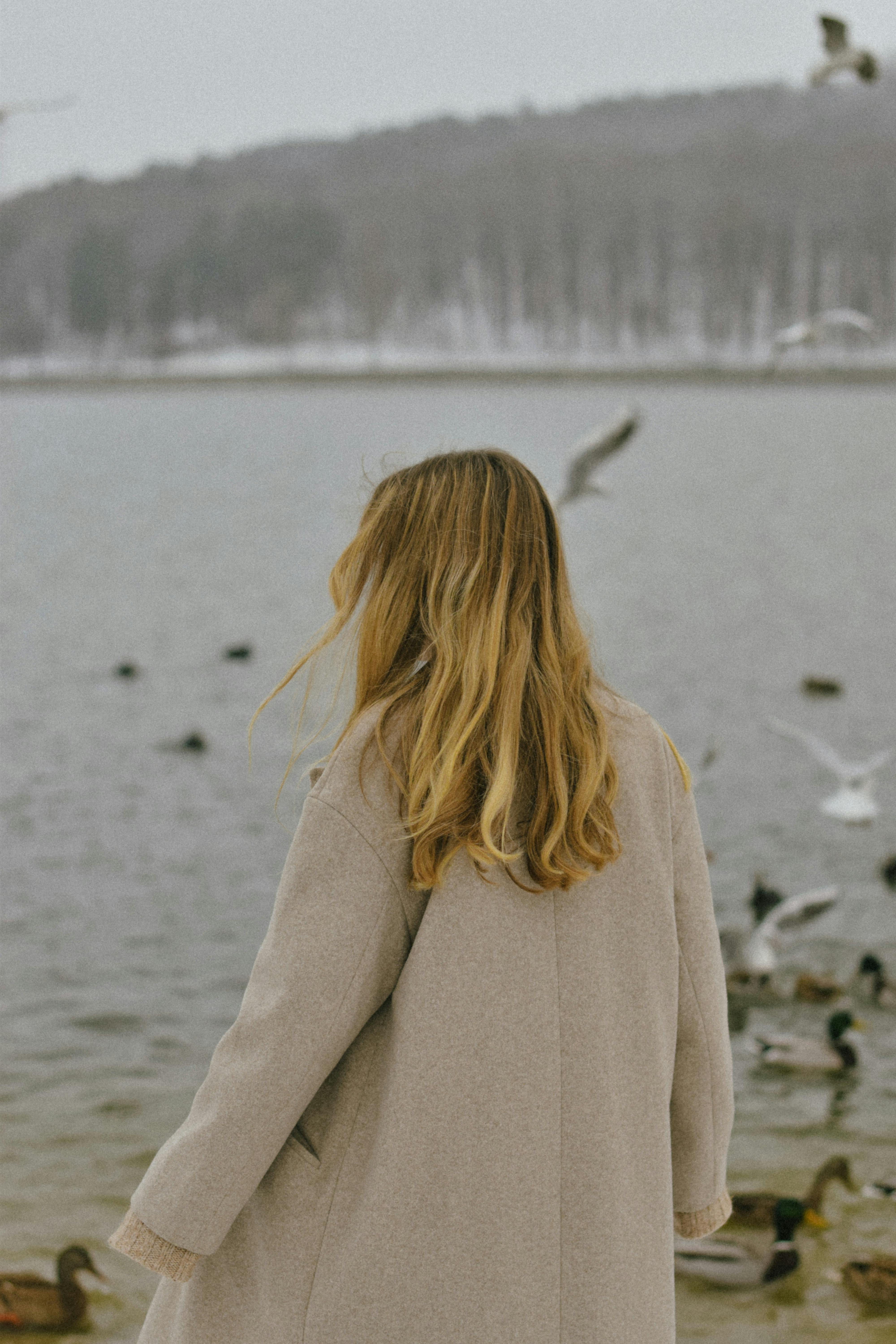 Blonde woman in winter coat by the lakeside with ducks and seagulls flying around.