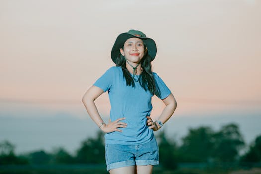 A smiling woman in casual attire poses confidently with hands on hips at sunset.