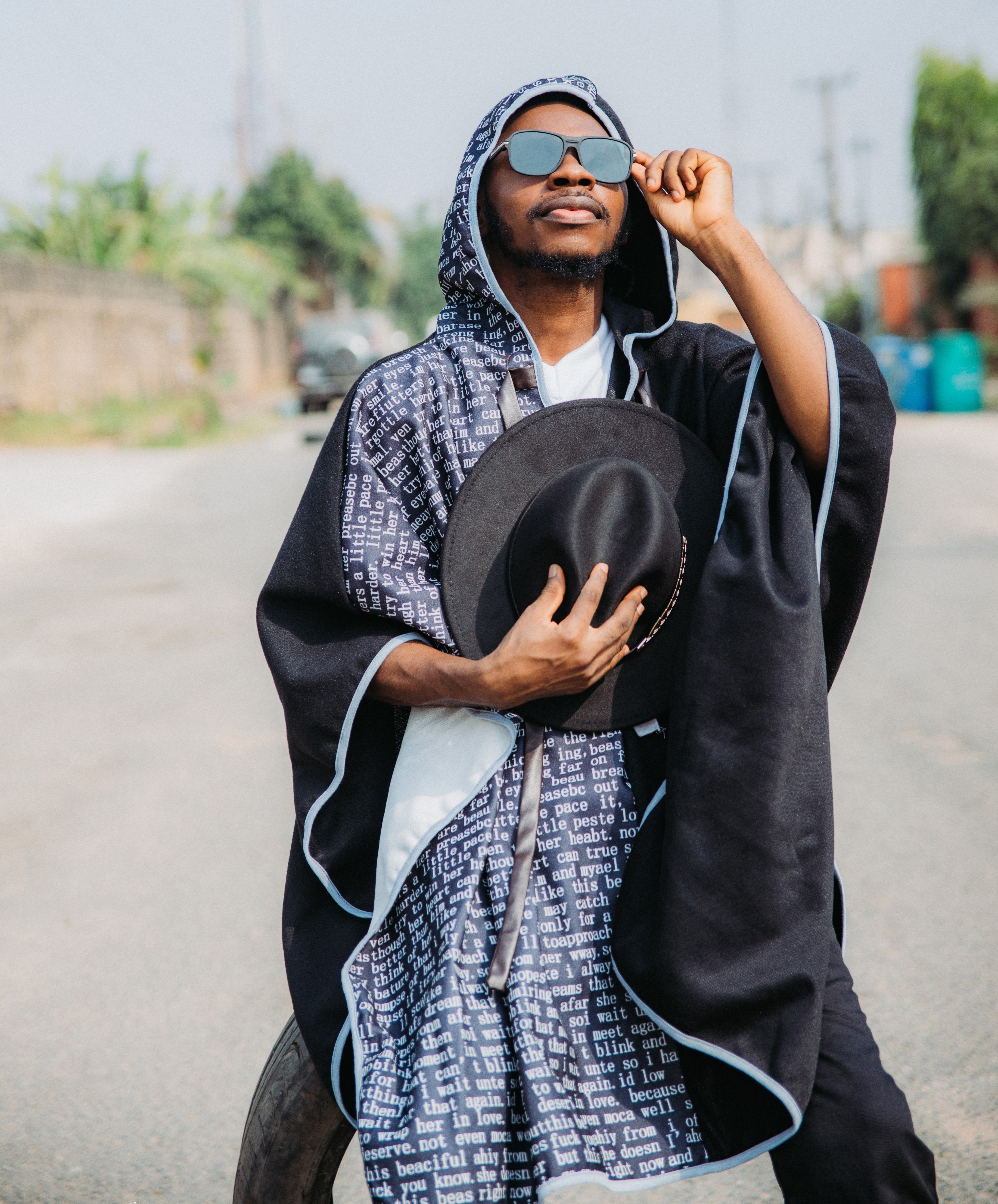 Fashionable man in a hoodie holding a hat, wearing sunglasses on a sunny street.