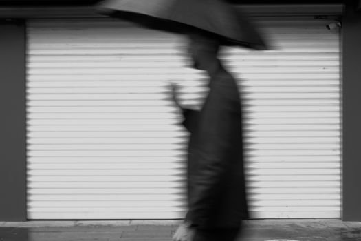 Black and white photo of a blurred man with umbrella in front of a shuttered storefront.