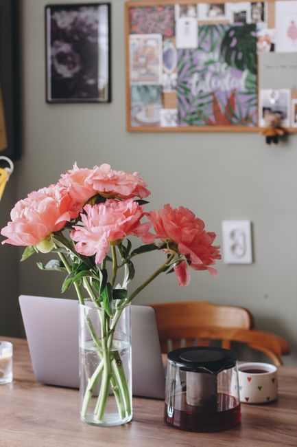 Pink flowers in a glass vase sitting on a wooden table beside a laptop and a coffee maker.