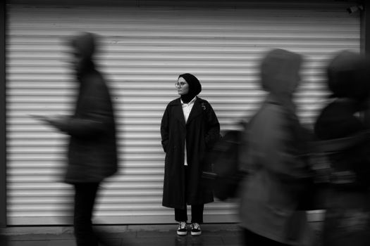 A black and white photo of a woman in a hijab standing still while people walk past her in an urban setting.