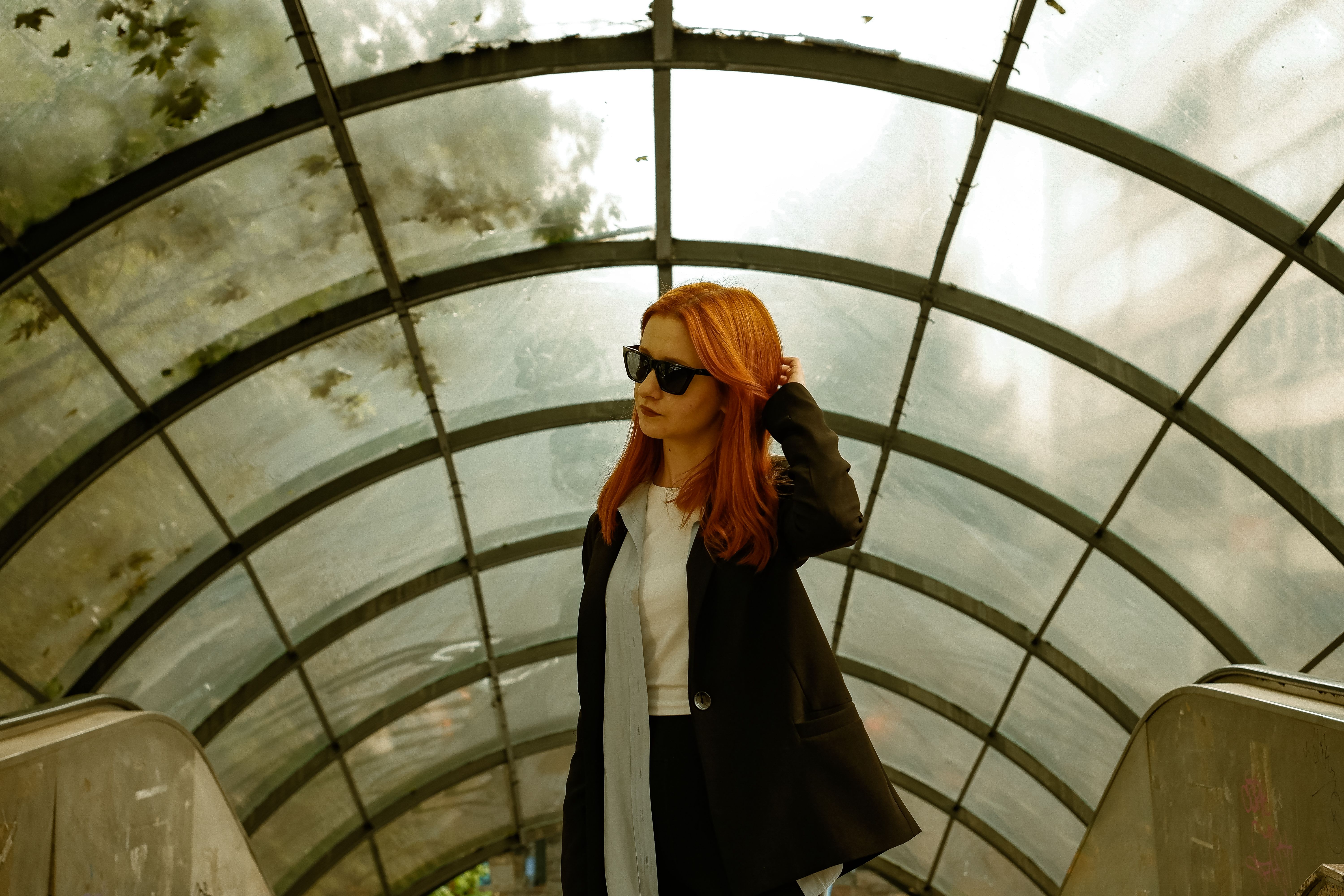 Redhead woman in sunglasses on an indoor escalator with glass canopy.