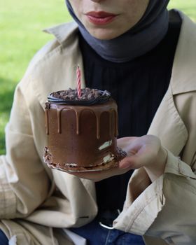 Woman holding a chocolate birthday cake with a candle in a park.