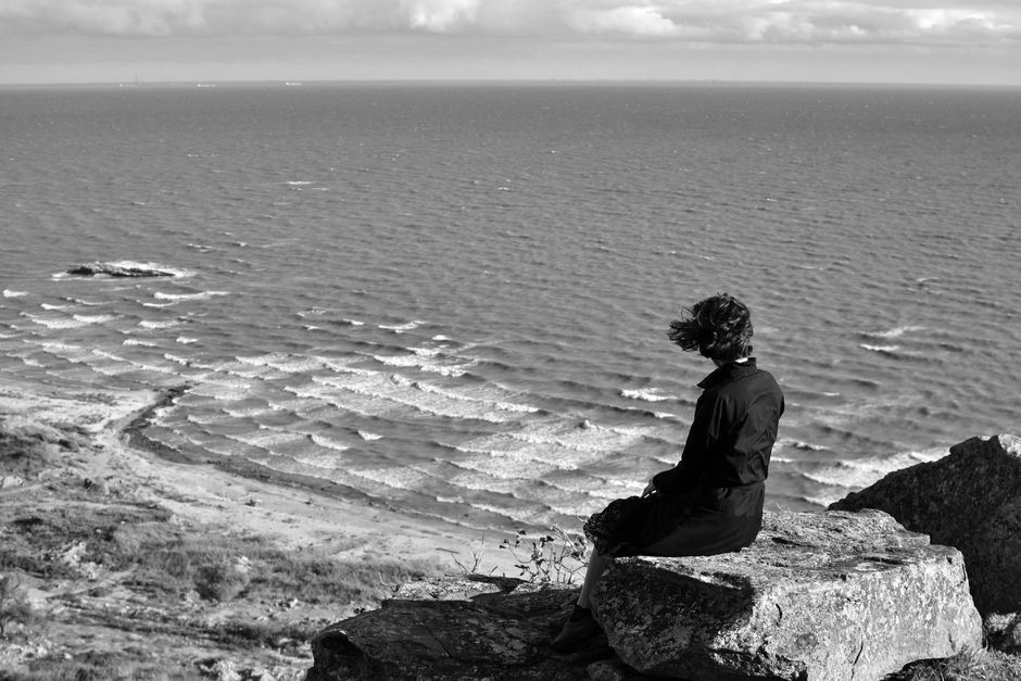 A woman sits on a cliff, gazing at the windy ocean. Black and white photography captures the scene.