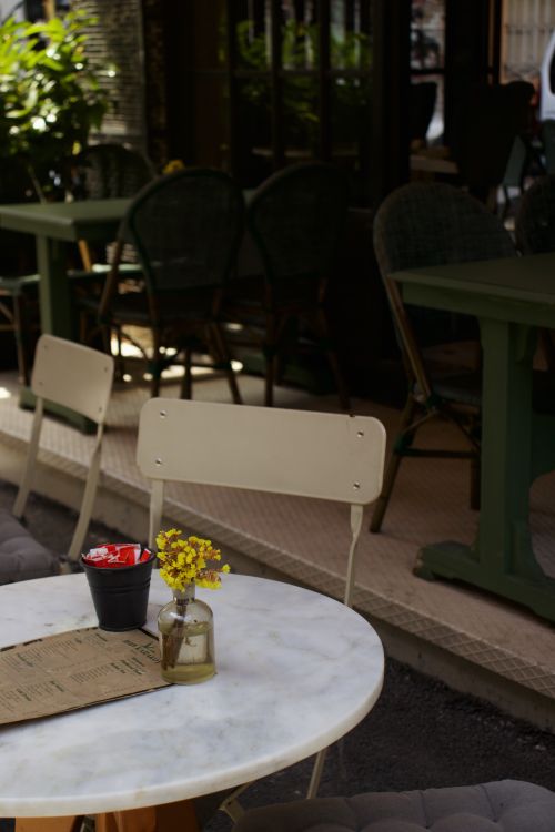 Free Cozy sidewalk cafe with tables, chairs, and yellow flowers in a vase for a serene dining experience. Stock Photo