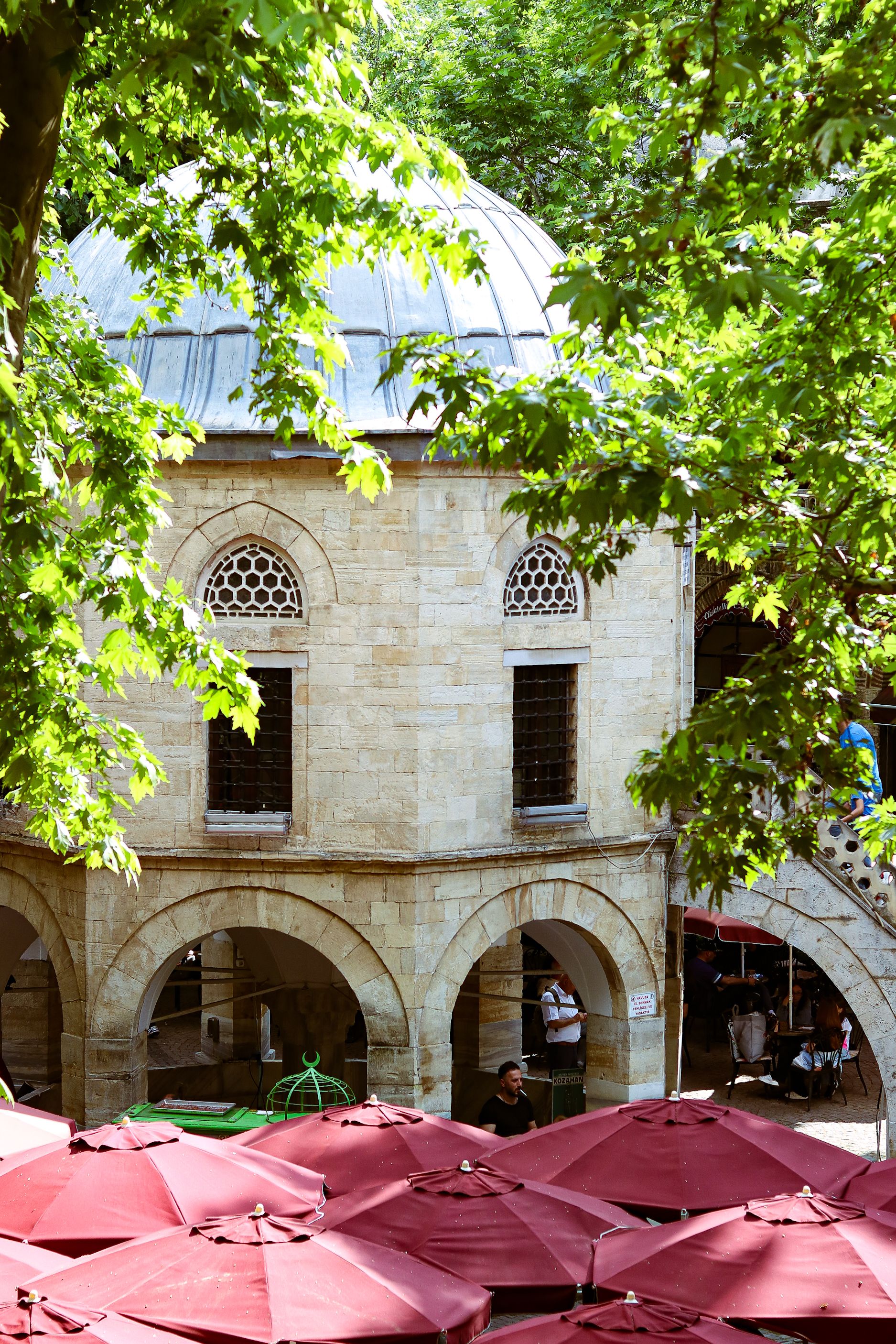 Explore the historic Koza Han in Bursa, Türkiye, surrounded by vibrant greenery and red umbrellas.