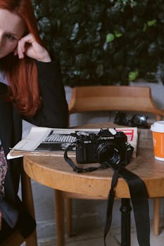 A vintage Petri camera and magazine on a café table with a woman seated nearby. Cozy and retro vibe.