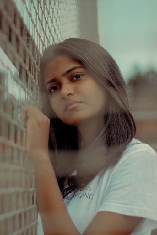 Free A young South Asian woman in a white t-shirt leans on a netting fence outdoors, looking pensive. Stock Photo