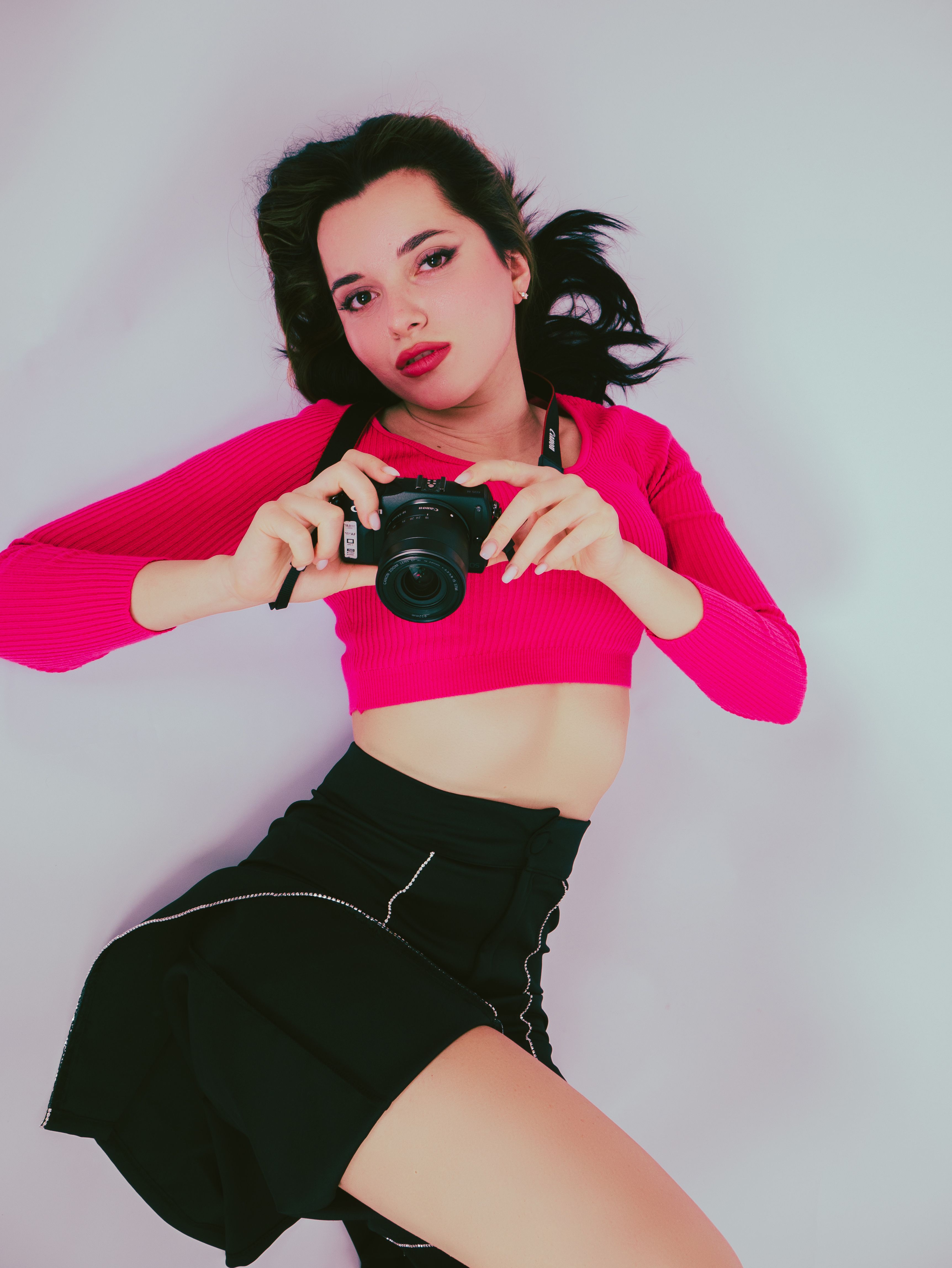 Portrait of a fashionable woman with black hair and a camera, wearing a pink top and black skirt.