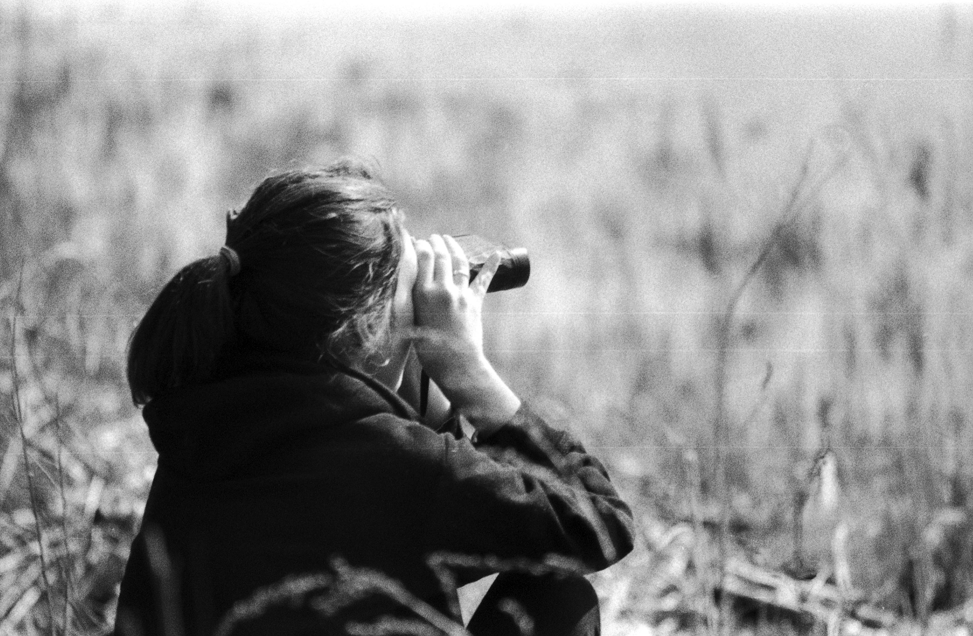 Black and white photo of a woman sitting in a meadow, using binoculars.