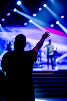 Silhouette of a man raising his hand at an energetic live concert indoors with vibrant stage lighting.