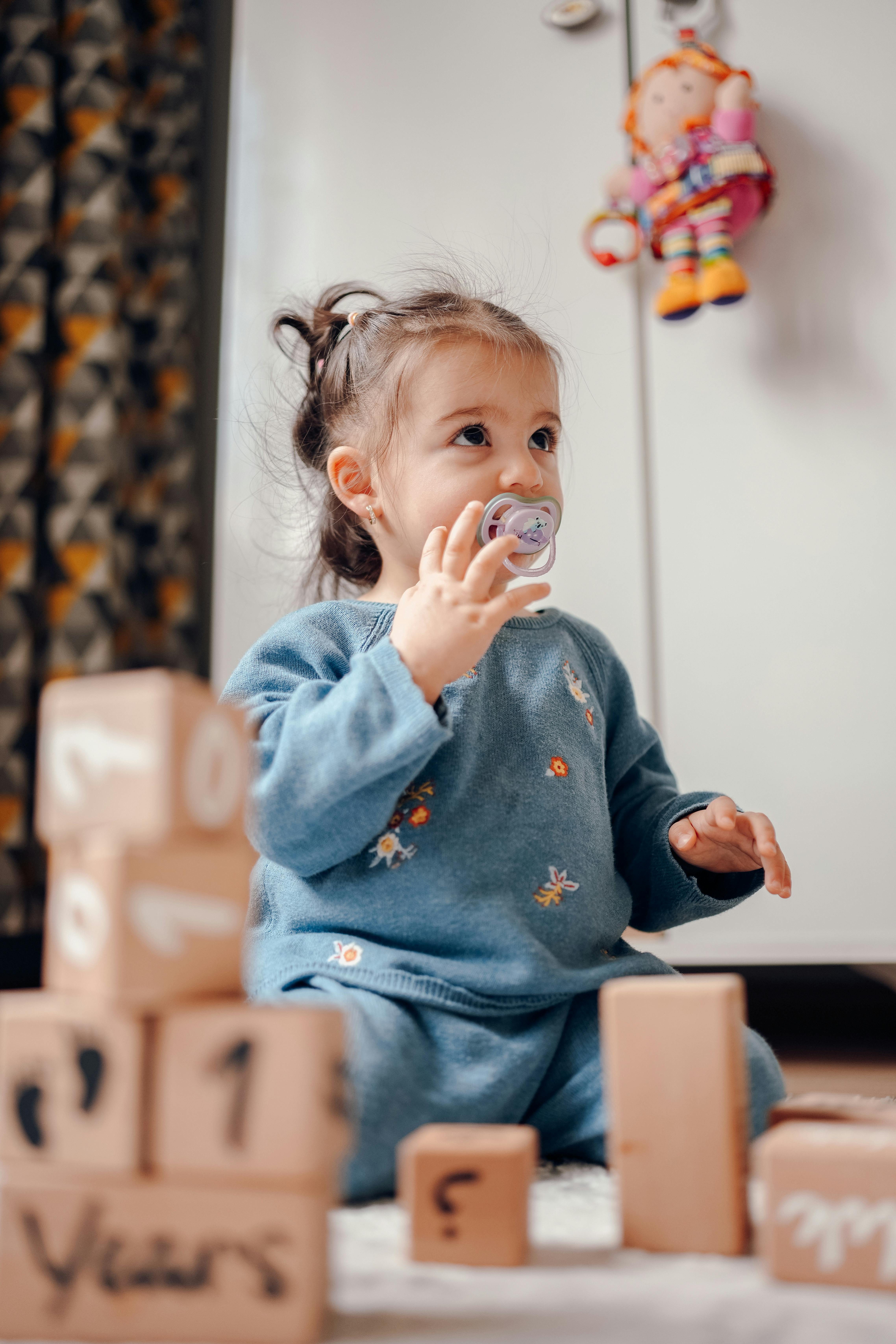 A cute child sitting indoors, playing with alphabet blocks and pacifier in a cozy room.
