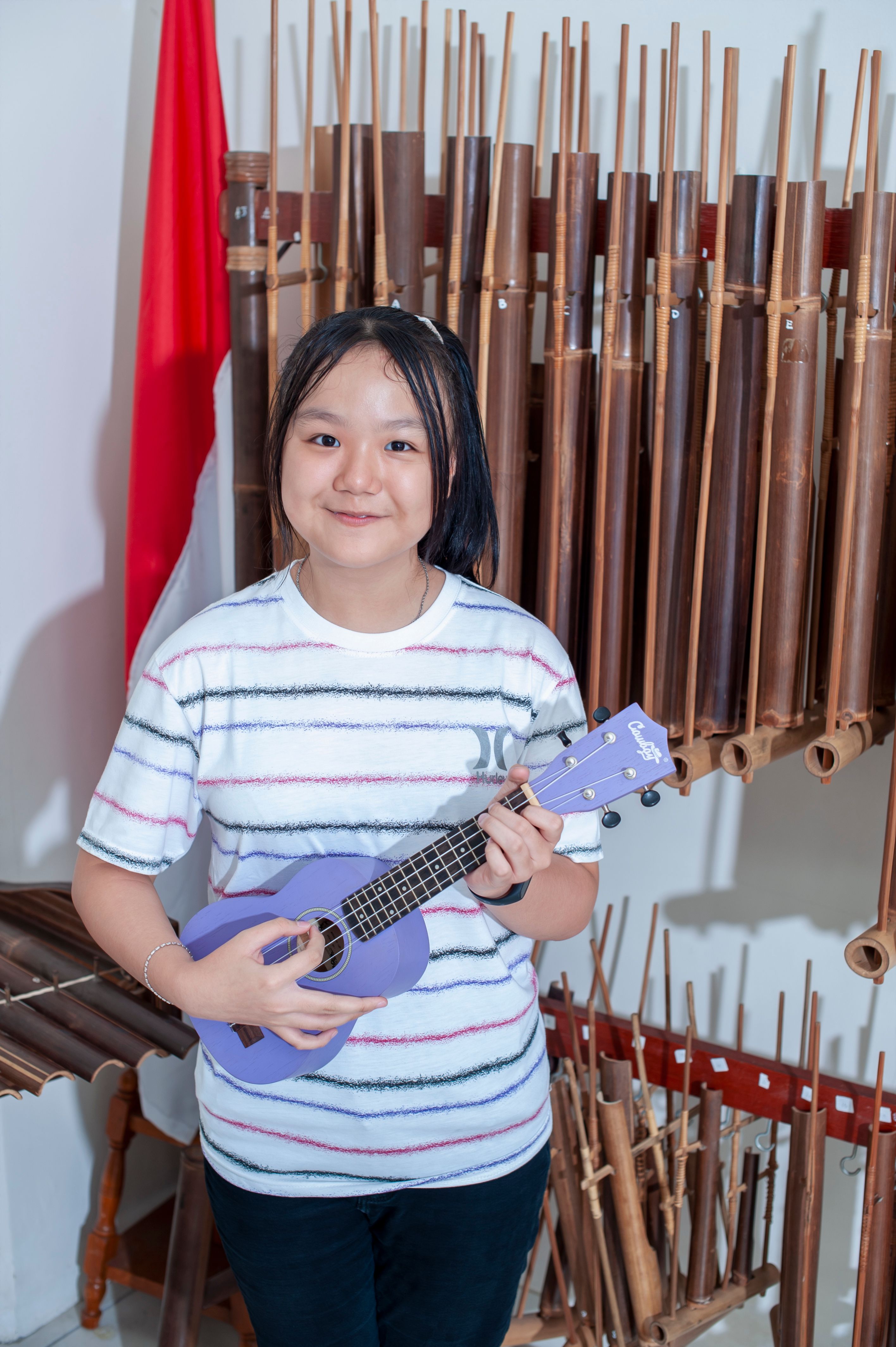 Smiling girl playing ukulele in front of traditional Indonesian bamboo instruments.
