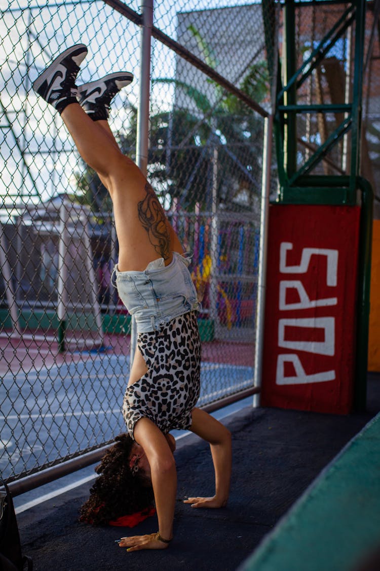 Young Fit Woman Making A Headstand By A Mesh Fence On A Sports Ground