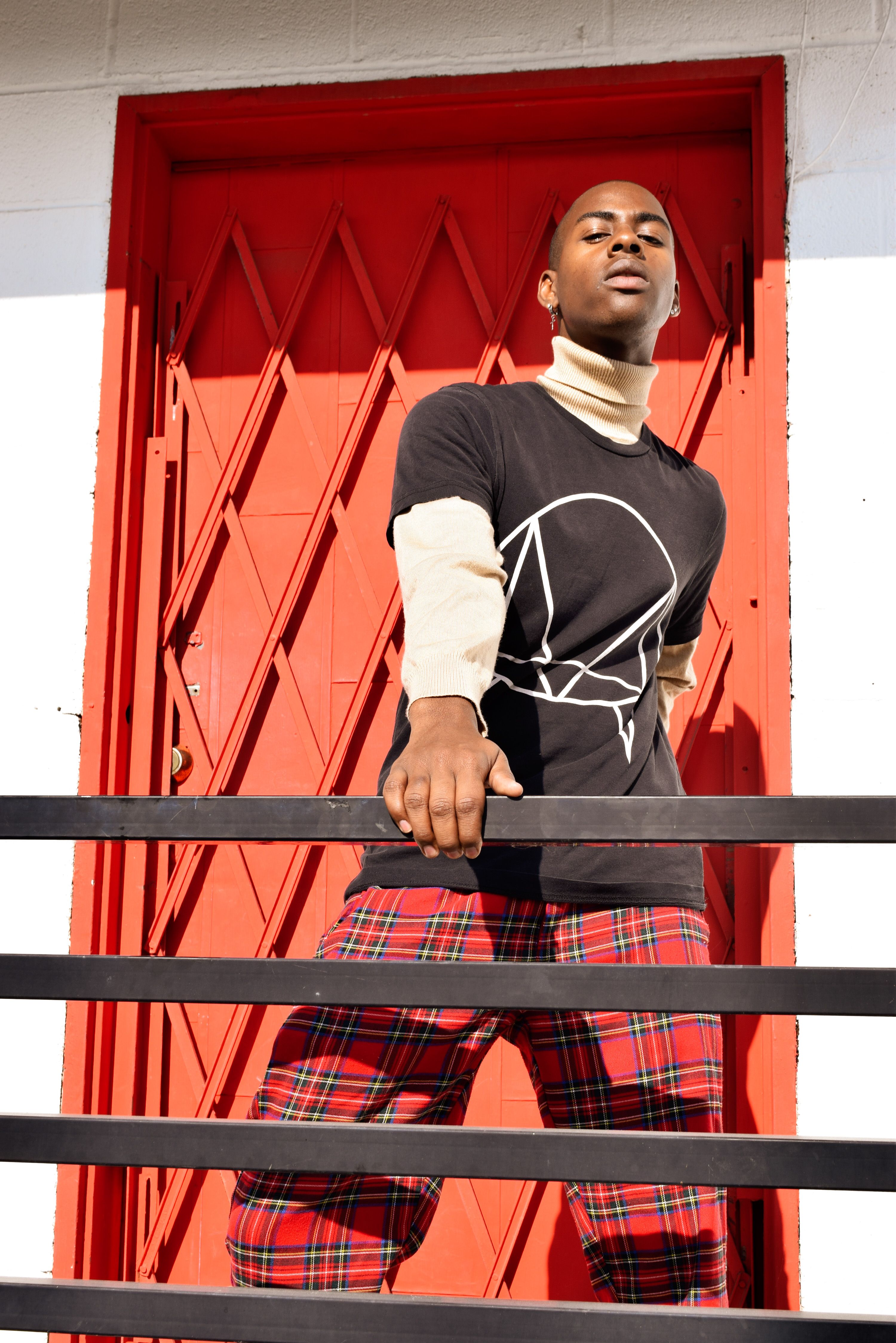 Fashionable young man in plaid pants and black t-shirt posing against a red geometric door.
