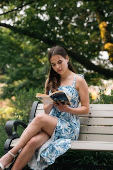 A young woman in a floral dress enjoys a book on a sunny day in a serene park setting.