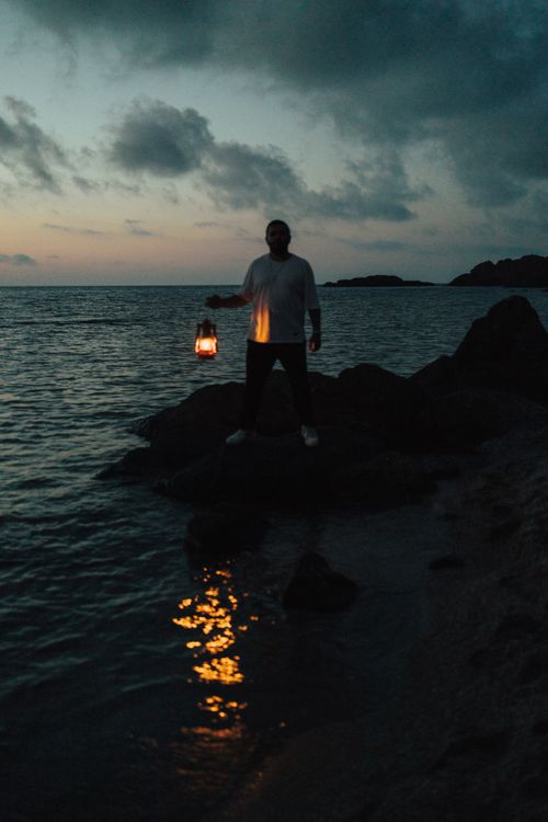 Free A person stands on rocks by the sea with a lantern at sunset, casting a warm reflection on the water. Stock Photo
