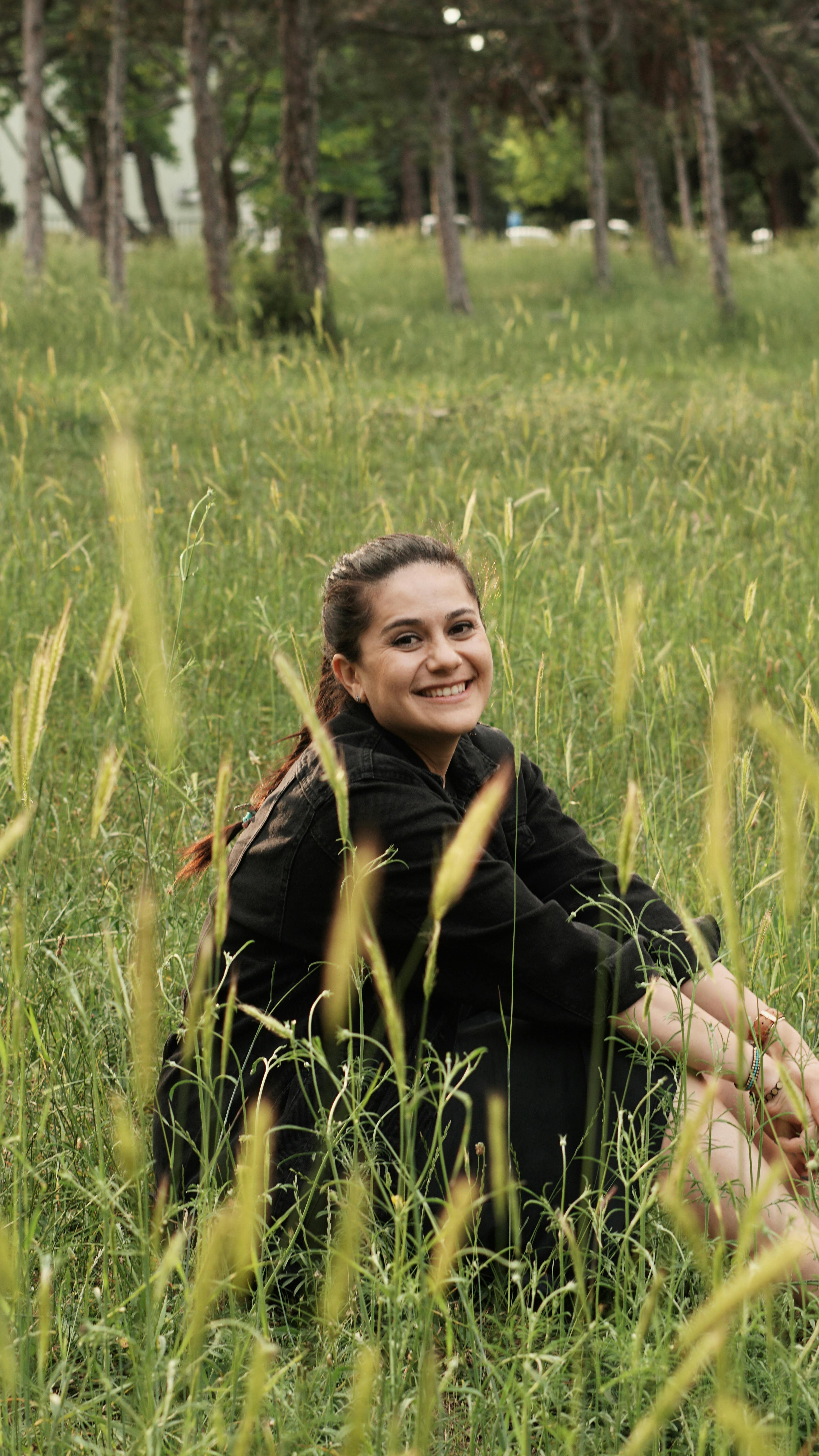 A happy woman in a black outfit sitting in a sunlit meadow surrounded by tall grass, exuding relaxation and joy.