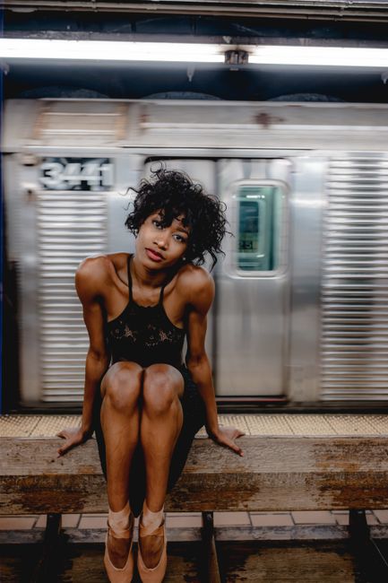 A young ballerina sitting in a subway station with a moving train in the background.