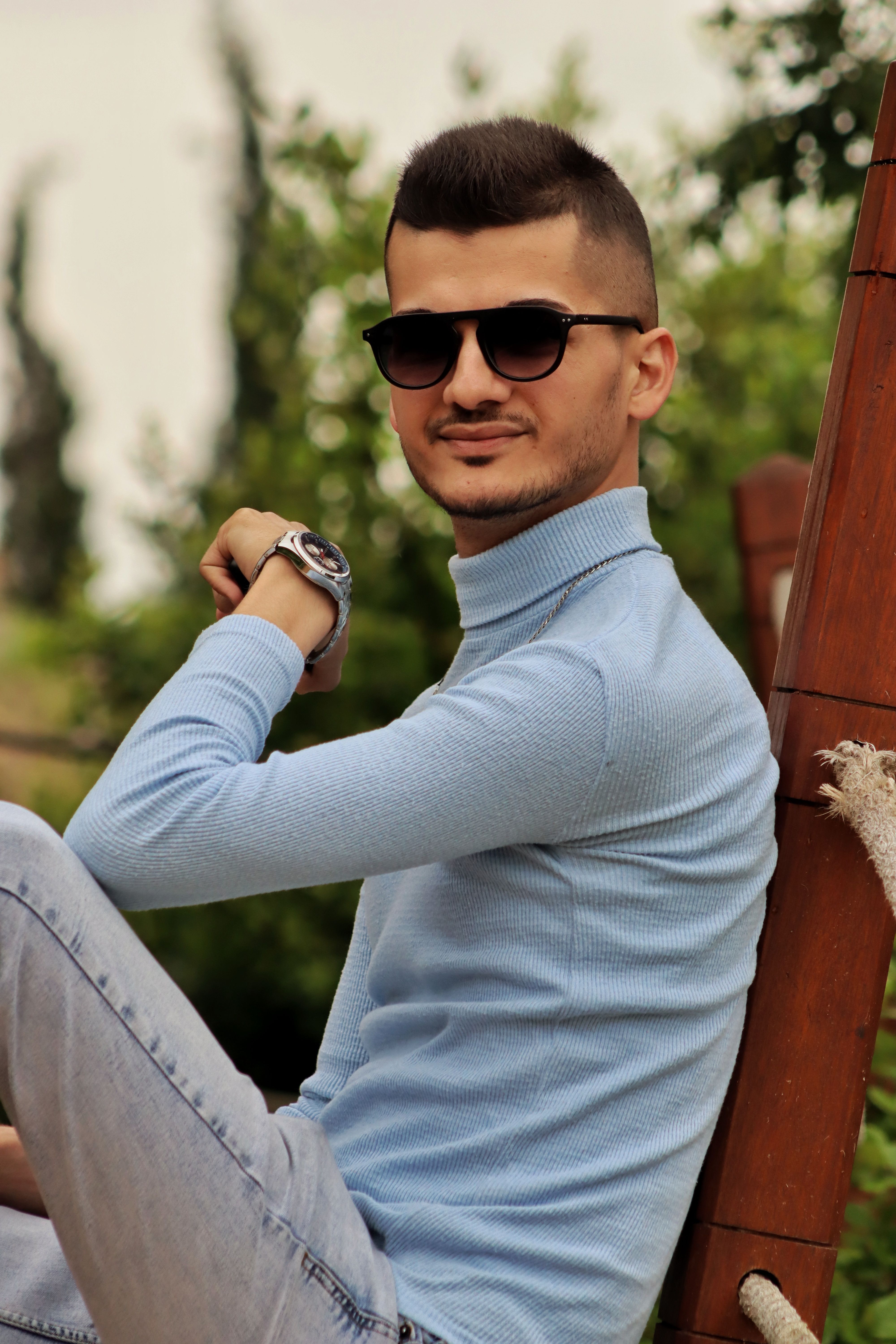 Young man in sunglasses and turtleneck sitting outdoors, showcasing casual style.
