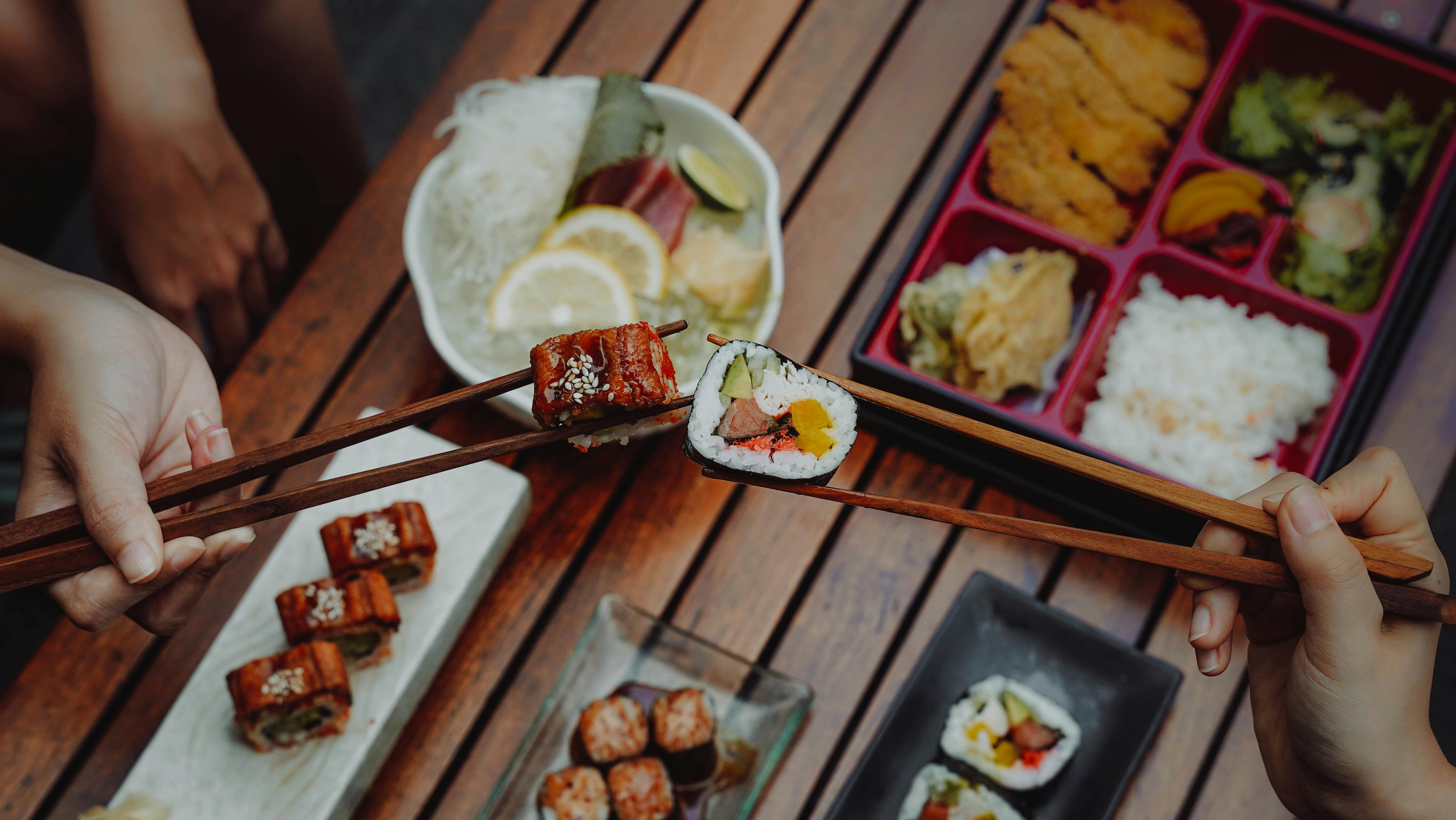Close-up of hands holding sushi with chopsticks, showcasing Japanese cuisine over a wooden table.