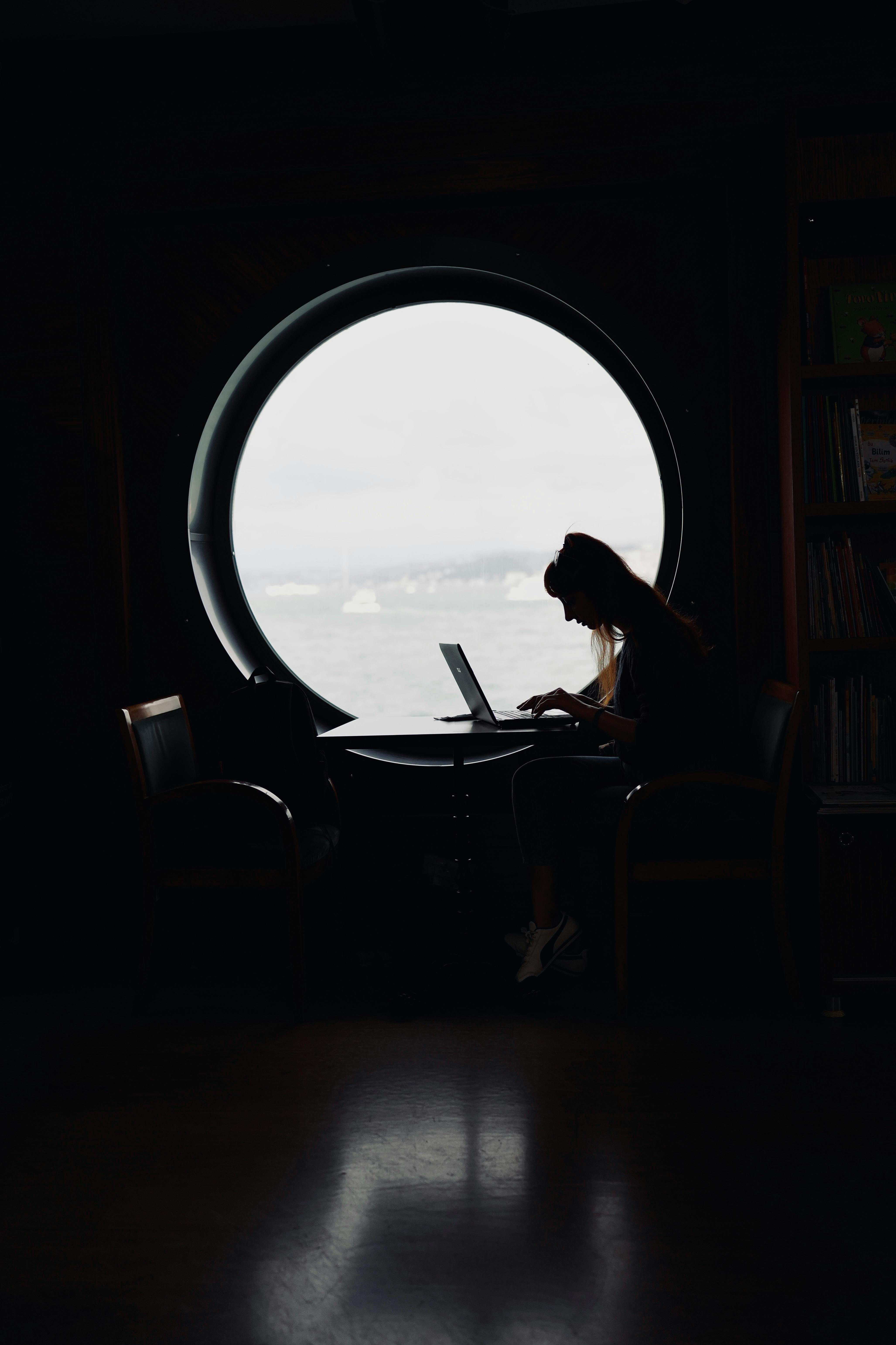A woman sitting by a porthole window working on a laptop in a dimly lit room, creating a silhouette effect.