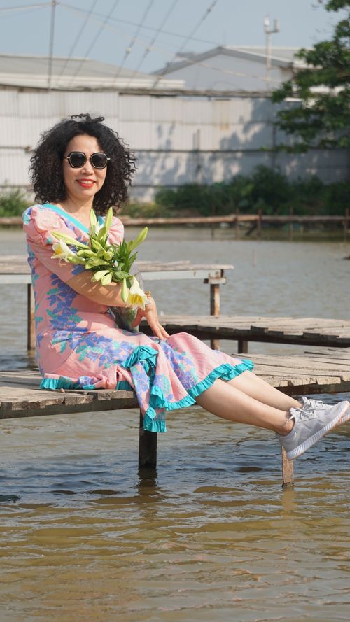 Free A joyful woman in a vibrant dress sits on a pier over water, holding flowers and smiling under the sun. Stock Photo