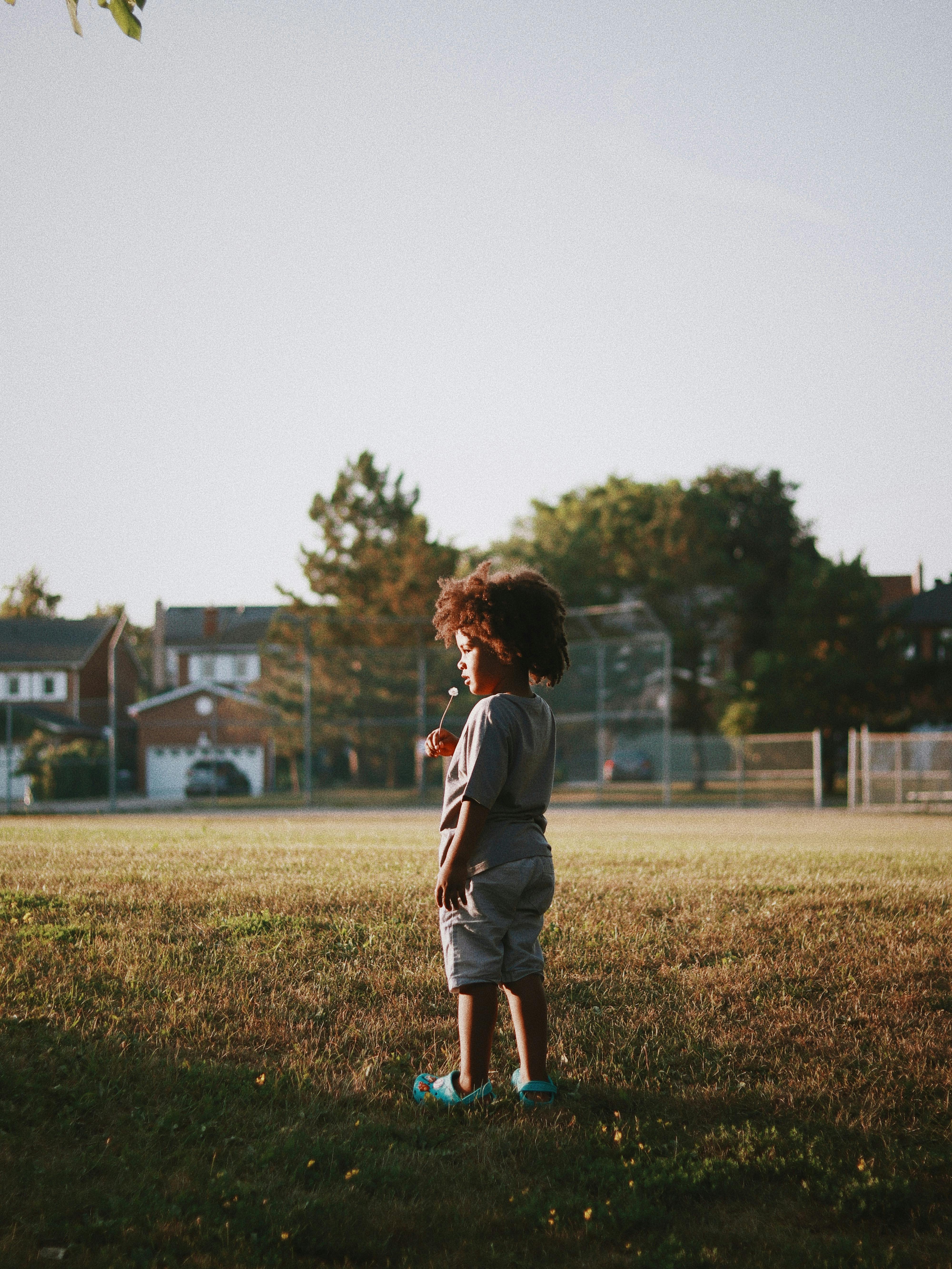 A young child with curly hair stands in a grassy park with houses in the background.