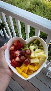 Heart-shaped bowl of fresh fruits held on a porch, evoking summer vibes.