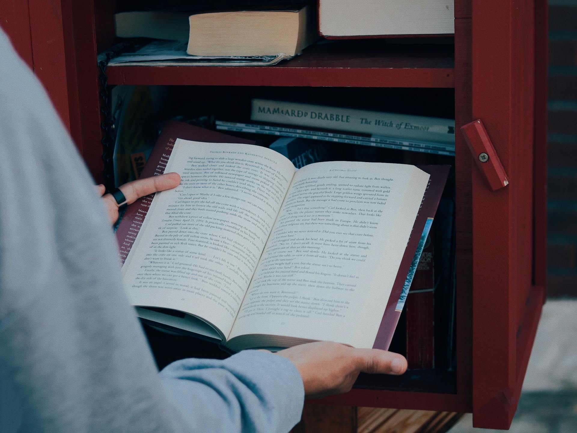 A person reads a book outside at a small free library, books visible on a wooden shelf.