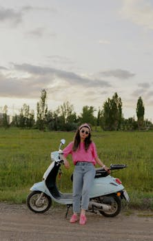 A woman in sunglasses and pink top stands by a scooter in a green countryside field.