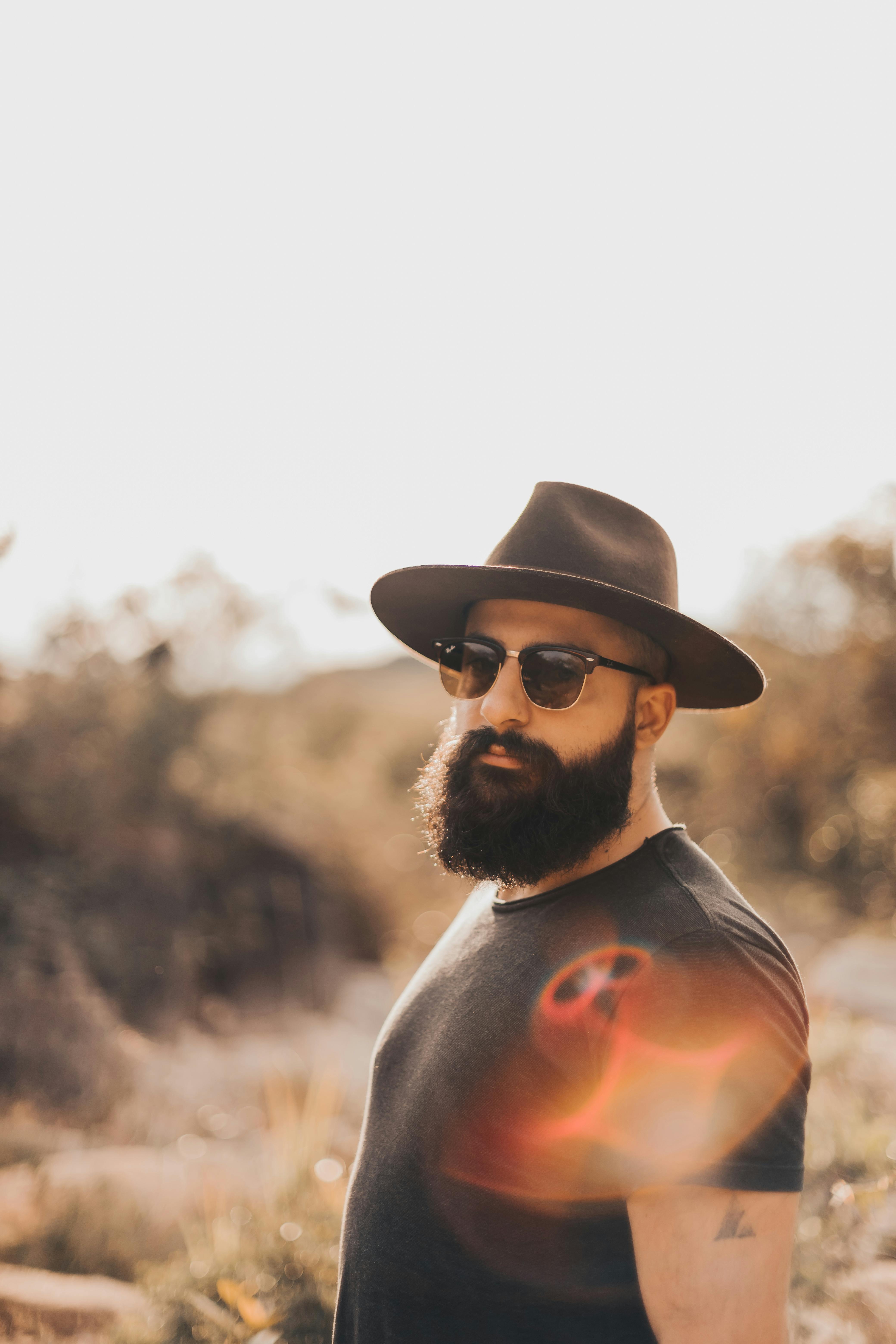 Portrait of a fashionable bearded man wearing a hat and sunglasses in an outdoor setting.