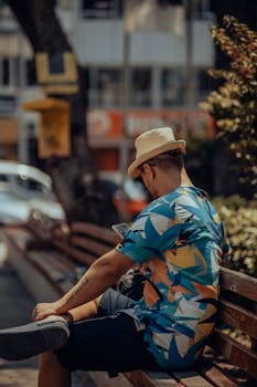 A man wearing a hat relaxes on a bench in a city, using his smartphone during summer.