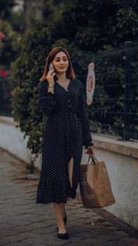Young woman in a polka-dot dress talking on the phone while walking with a shopping bag on an urban sidewalk.