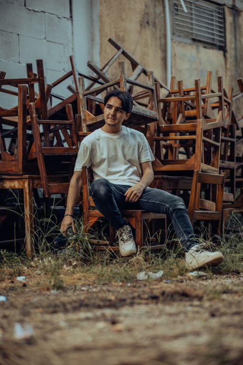 Free Young man sitting casually by a pile of wooden chairs outdoors, wearing jeans and sneakers. Stock Photo