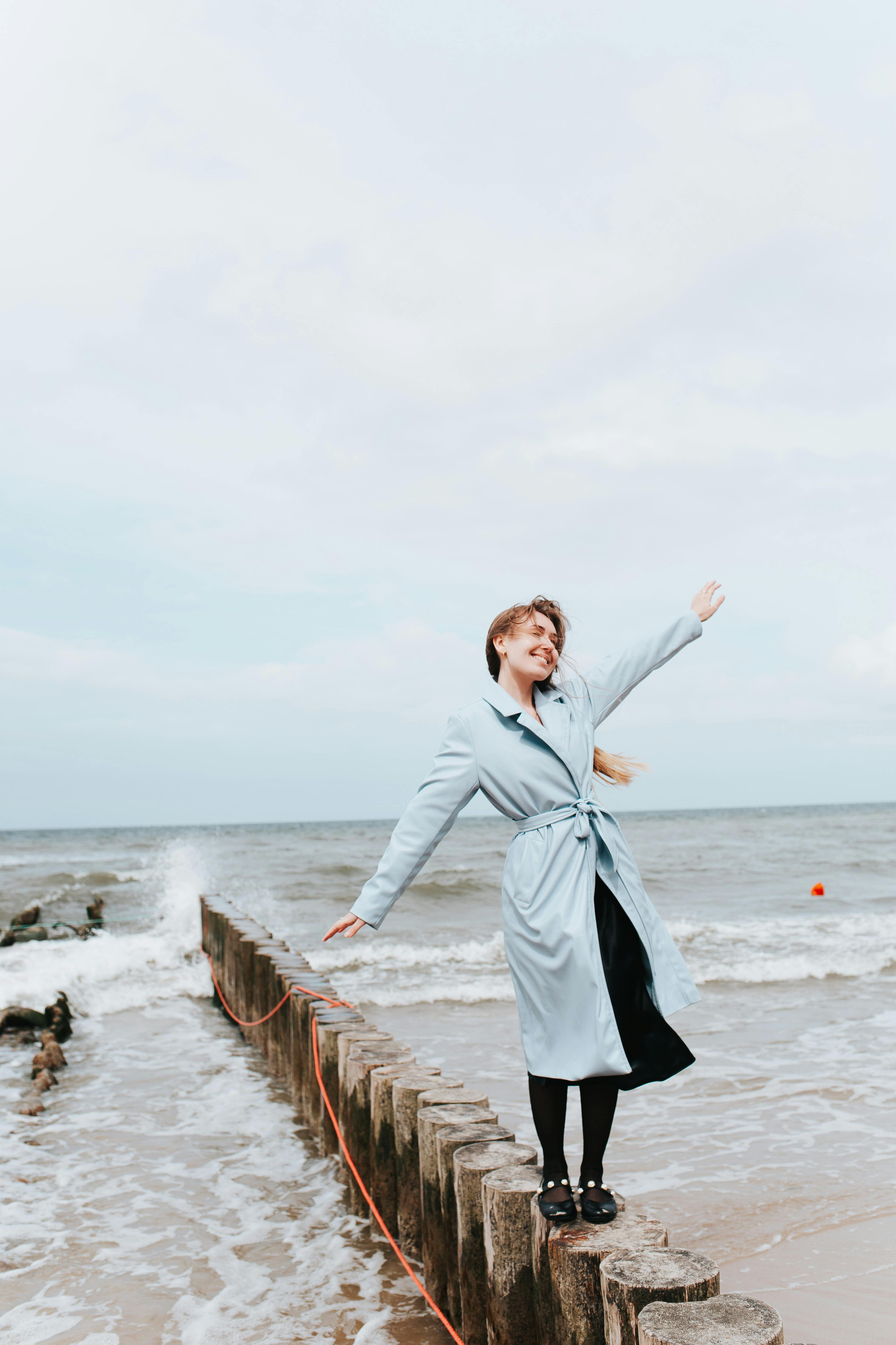 Woman in blue coat smiling with arms outstretched, standing on the shore.