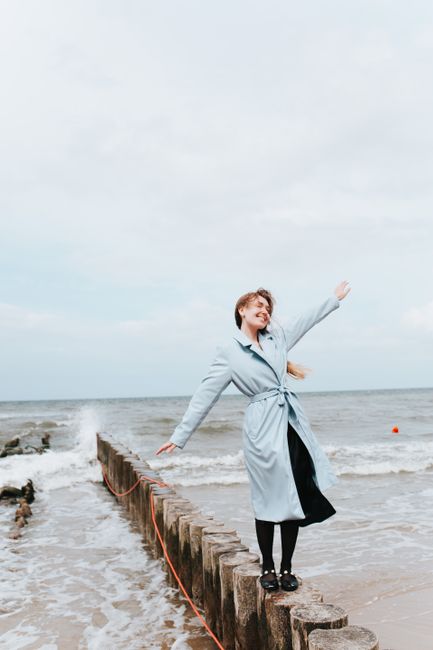 Woman in blue coat smiling with arms outstretched, standing on the shore.