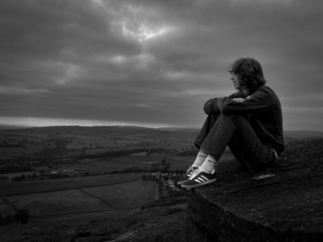 Black and white portrait of a man sitting on a cliff, looking over fields under a dramatic sky.