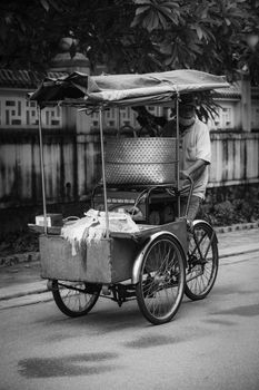 Black and white photo of a man pushing a bicycle rickshaw on a city street. Urban transportation.