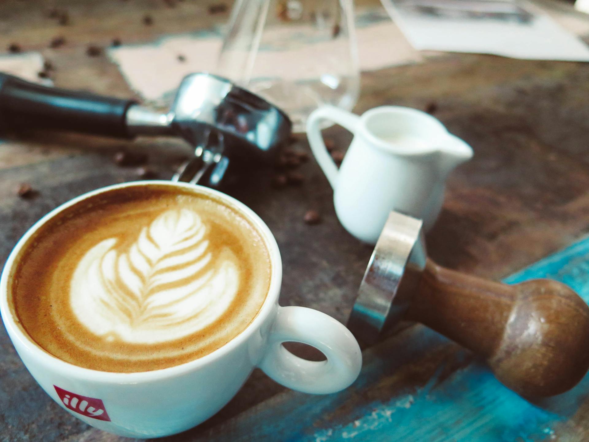 A latte with elegant latte art accompanied by a milk jug and coffee tamper on a rustic table.