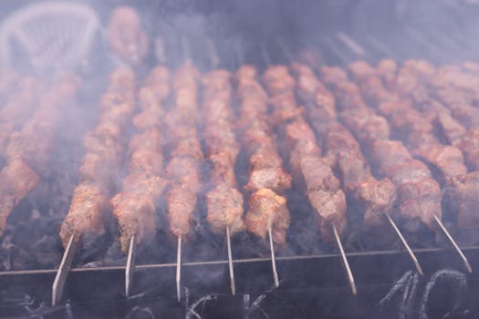 Close-up of well-seasoned skewered meats grilling with visible smoke on a barbecue grill.